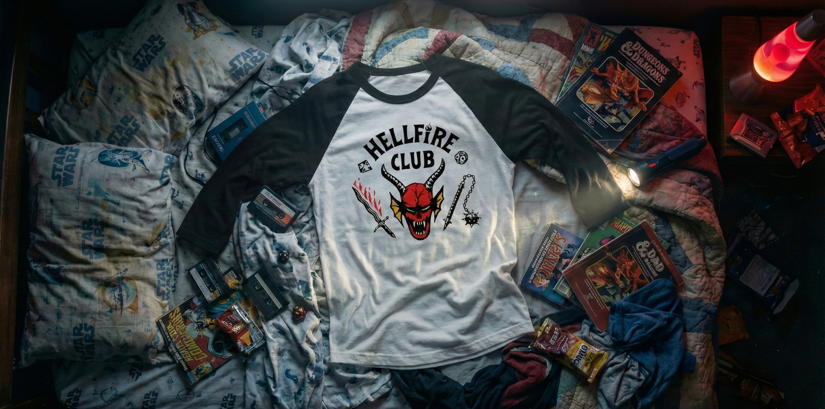 A white and black raglan t-shirt with a 'Hellfire Club' graphic from Star Wars, laid flat on a bed surrounded by Star Wars themed bedding, collectible cards, and a glowing lamp.